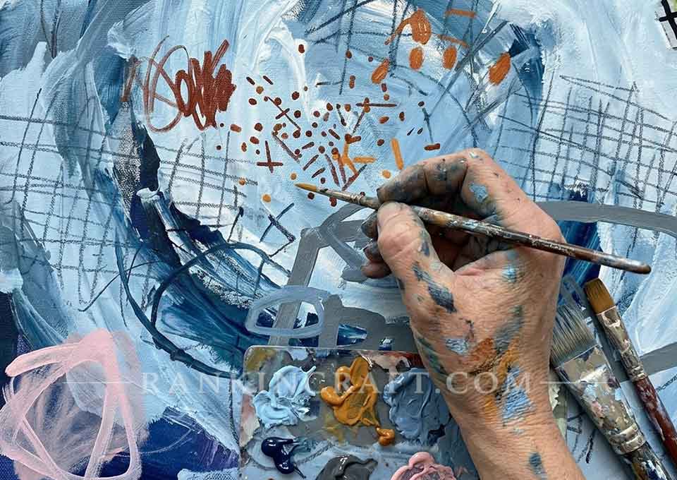 close-up of artist hand holding a paintbrush adding fine details to a blue abstract canvas